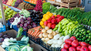 Colorful display of fresh fruits and vegetables at a New Jersey farmers market, including tomatoes, cucumbers, peppers, onions, carrots, and leafy greens.